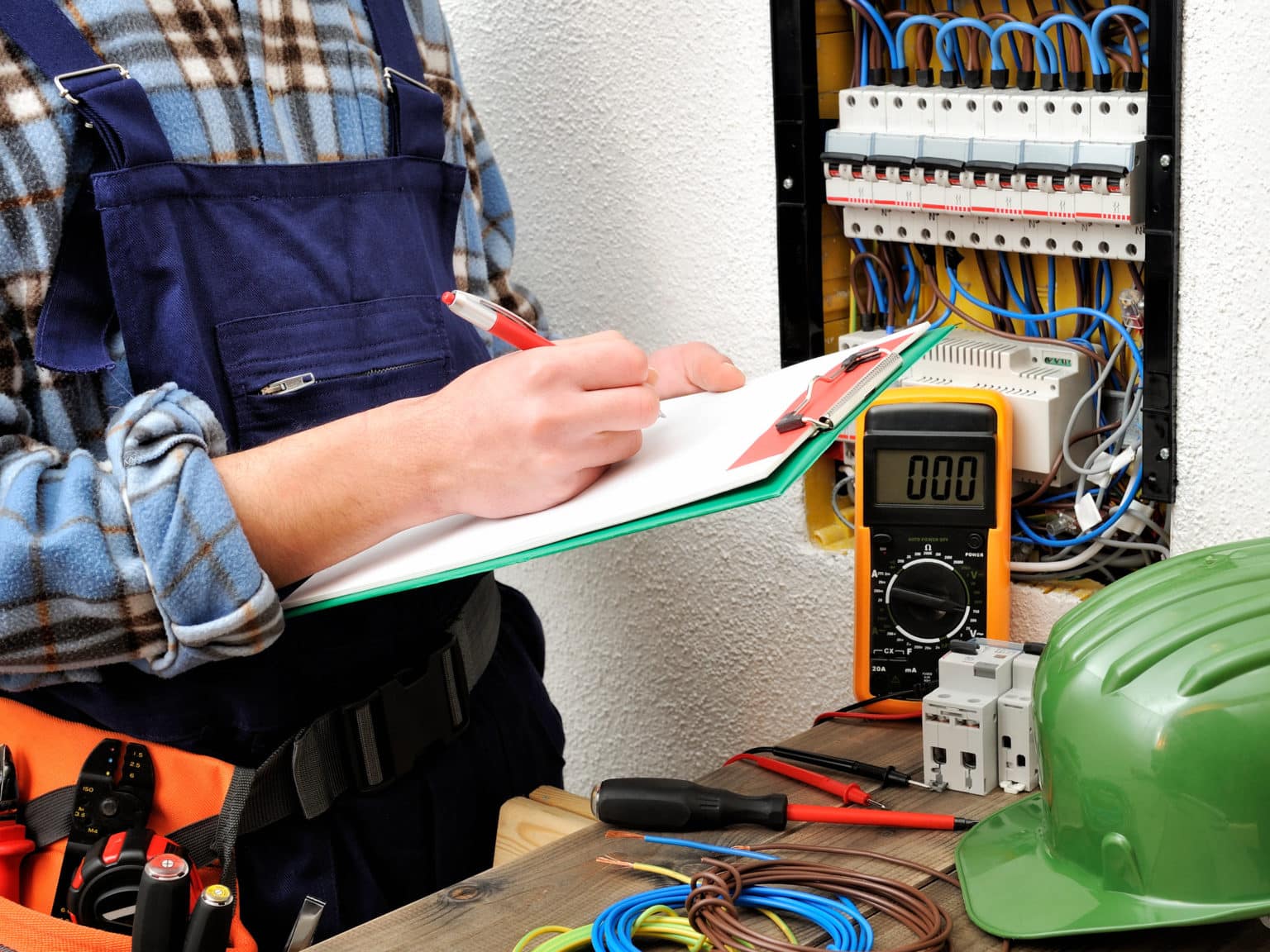 Electrician in work overalls writing on a clipboard while inspecting an electrical panel with tools and a multimeter nearby.