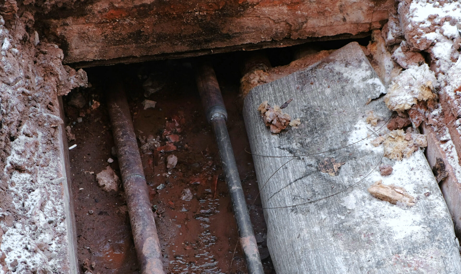 Old corroded metal pipes exposed underground during excavation, surrounded by soil, debris, and crumbling concrete.
