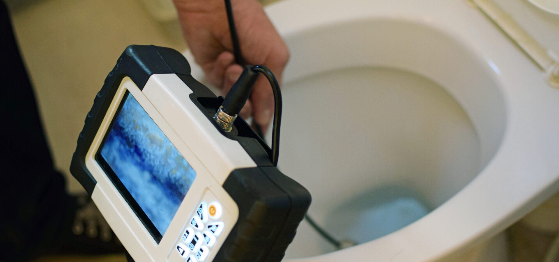 Plumber using a sewer inspection camera to examine the inside of a toilet drain, with the monitor showing a live feed of the pipe interior.