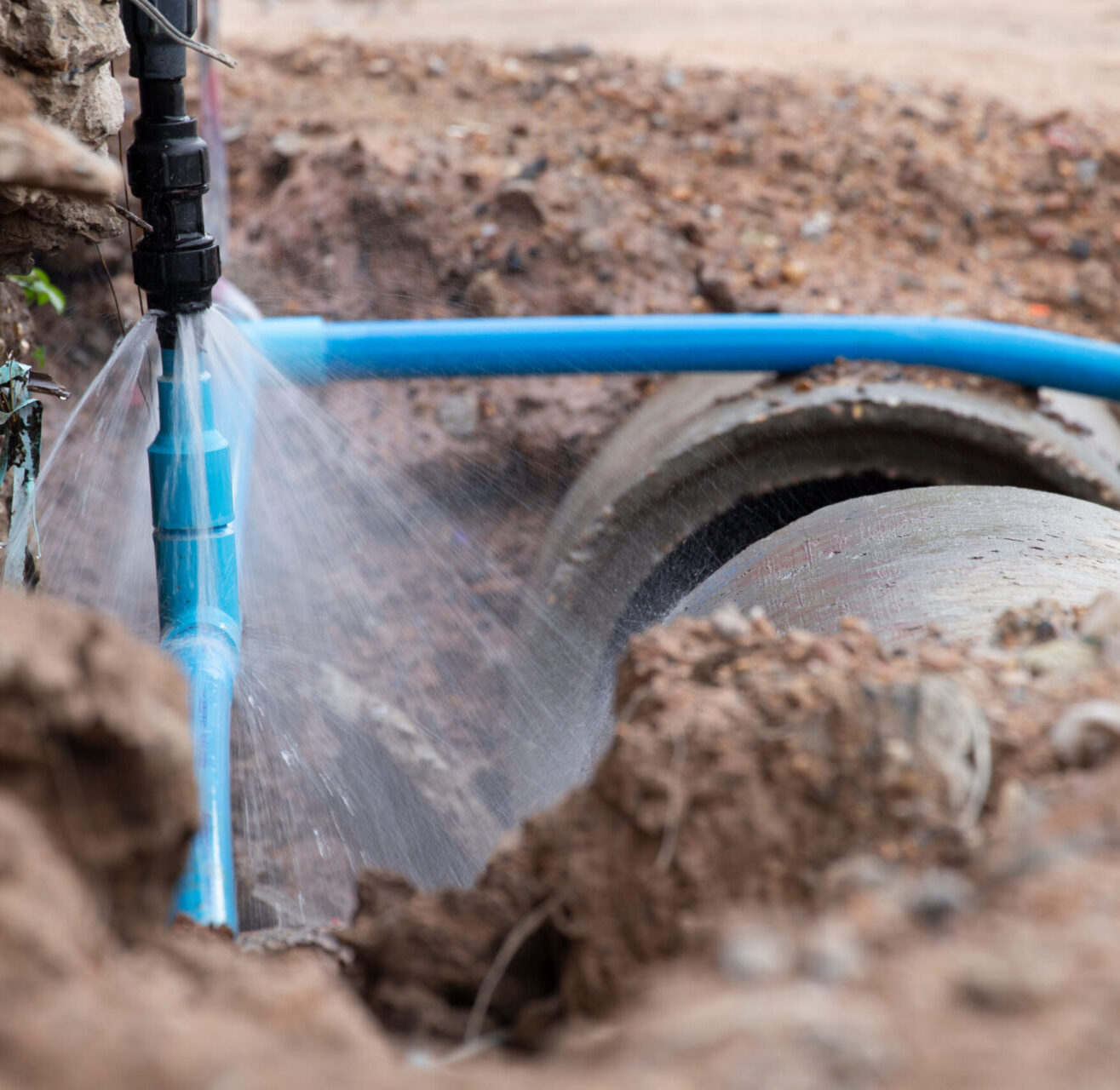 Broken underground blue water pipe leaking and spraying water in a dirt trench.