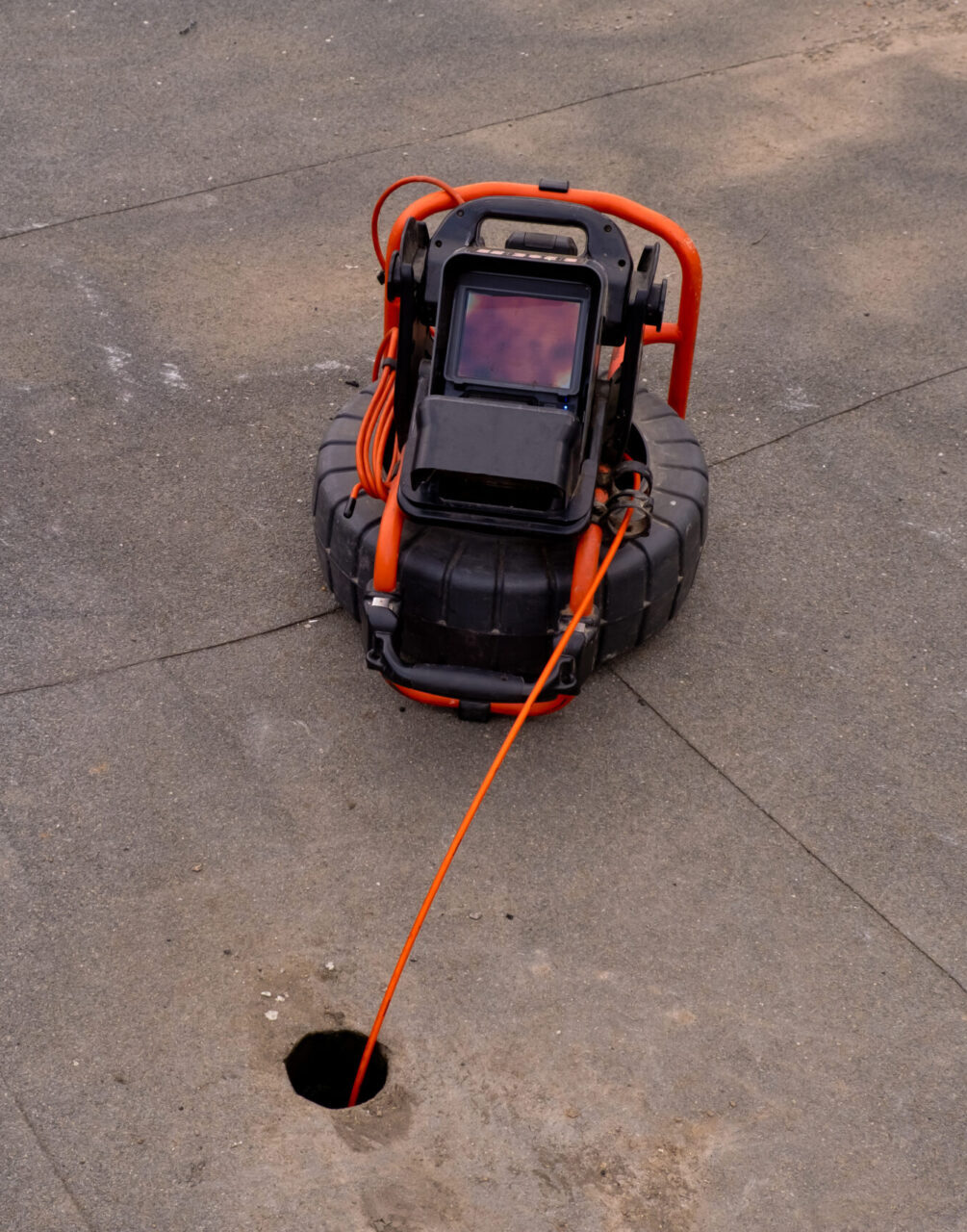 Sewer inspection camera with an orange cable inserted into a drain opening in concrete pavement.