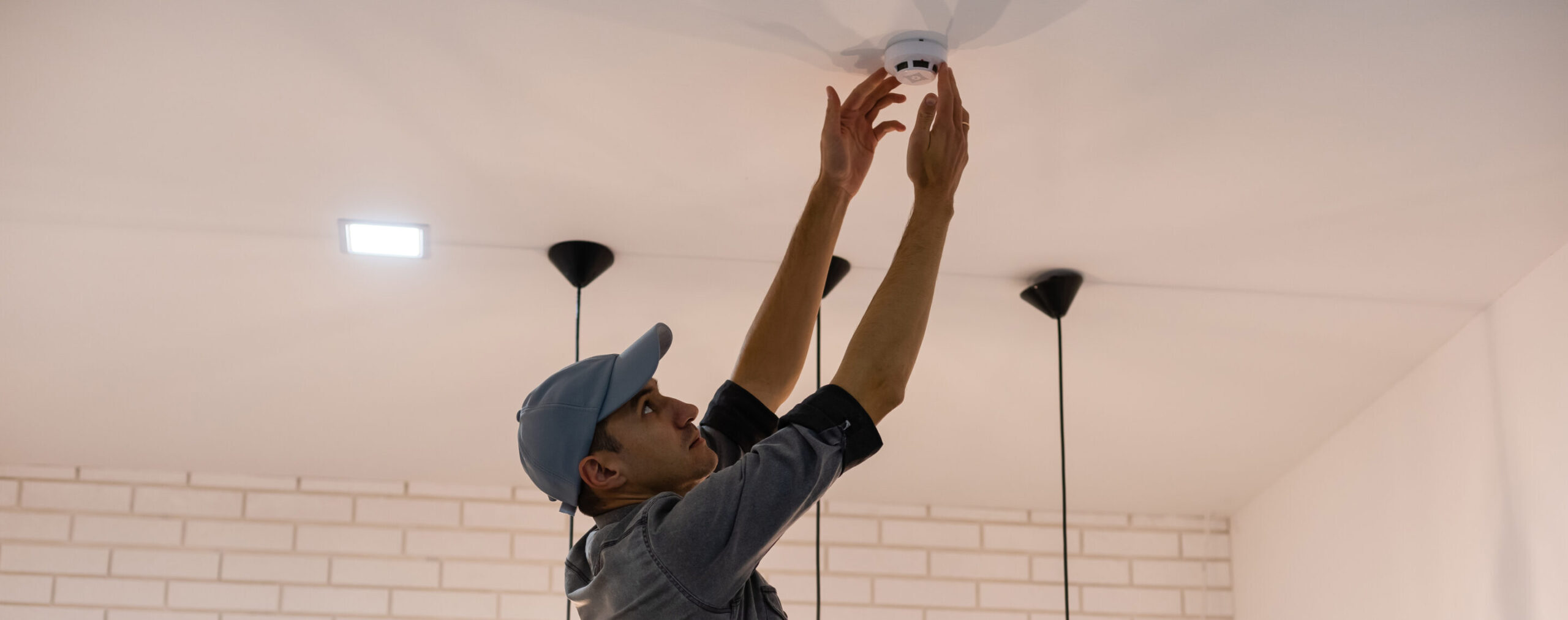 Man in a blue cap installing a smoke detector on a ceiling in a modern room.