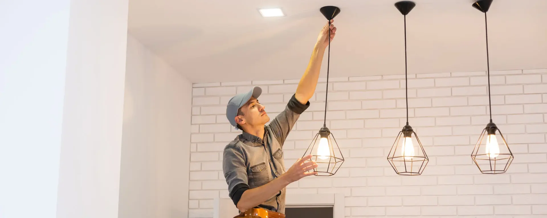 Electrician adjusting a hanging pendant light fixture in a modern interior with white brick walls.
