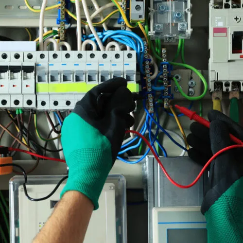 Electrician wearing green gloves working with wiring inside an electrical breaker panel.