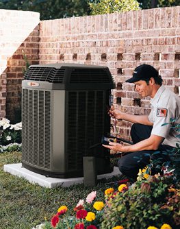 An HVAC technician kneels outside next to a large outdoor air conditioning unit, using tools to perform maintenance. The unit is placed on a concrete slab beside a brick wall, surrounded by a well-kept garden with colorful flowers.